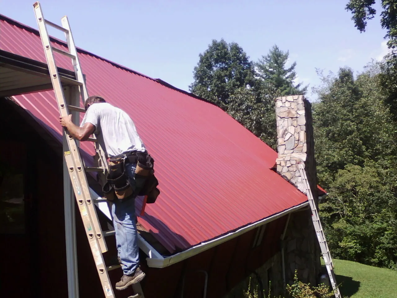 Roofer on a ladder beside a red metal roof and stone chimney — Soffit & Fascia Repair by Roofing Brothers in Amissville